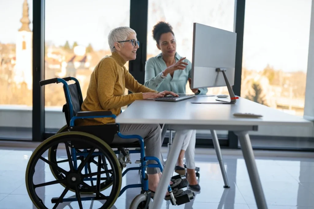 Disabled woman in wheelchair at work with colleague looking at computer together